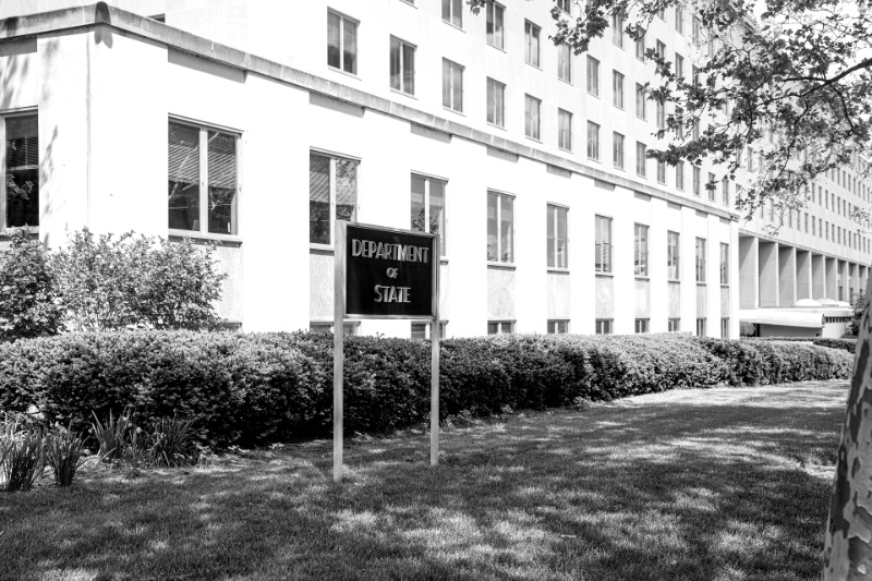 A Department of State sign on a lawn in front of the long pale stone Harry S. Truman Building in Washington, a clipped hedge along its facade and a tree in leaf at the right
