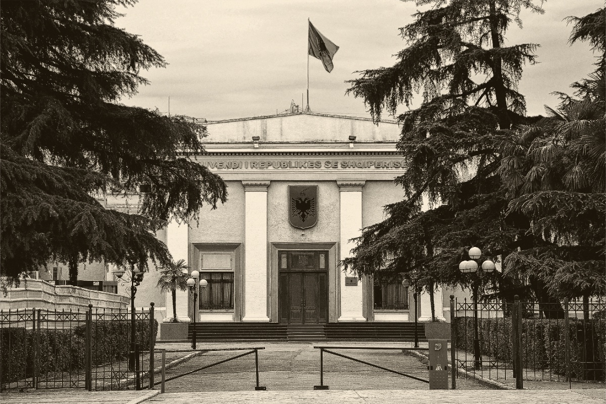 The National Assembly building in Tirana seen through its iron gates, a pale columned entrance with the Albanian flag on the roof and tall cedars on either side, in an ink and bone duotone
