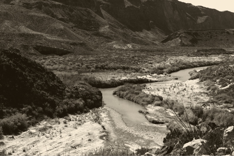 The Rio Grande winding between gravel bars and green scrub below dark desert hills in Big Bend Ranch State Park, Texas, seen from above, in a warm ink and bone duotone
