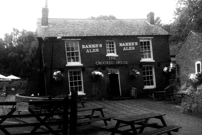 The Crooked House pub in Himley, a two-storey brick building leaning visibly to one side, signs reading Banks’s Ales and The Crooked House on its front, empty picnic benches on the paved yard, in grainy high-contrast black and white