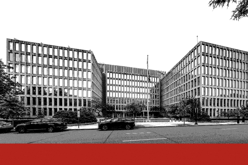 The rectilinear concrete and glass facade of the Theodore Roosevelt Building in Washington with flags outside, a deep band running across the foot of the frame