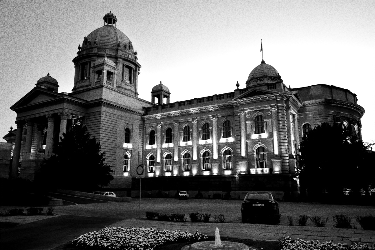 The House of the National Assembly in Belgrade at twilight, its domes dark against the sky and its windows lit, a fountain and flower beds in the foreground, in high contrast black and white with coarse grain