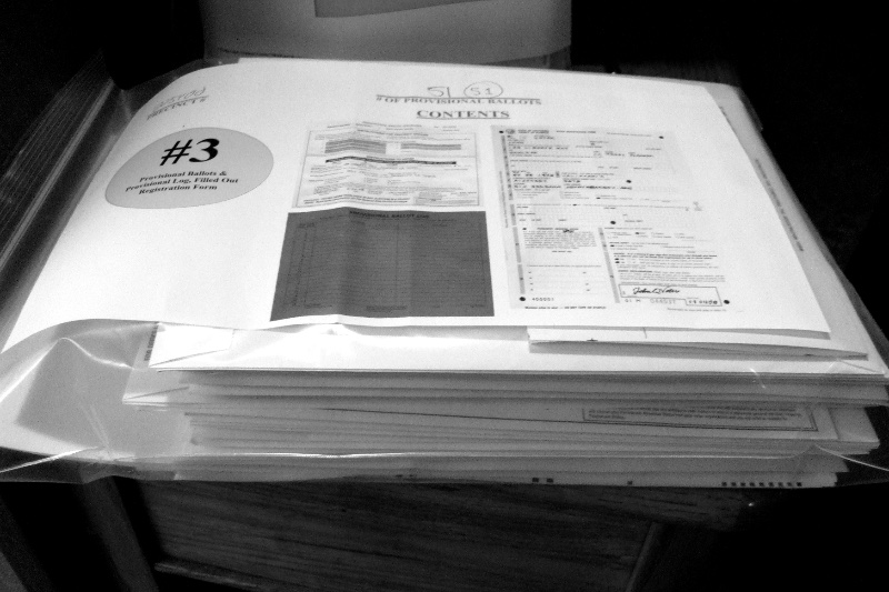 A clear plastic bag holding a stack of provisional ballot envelopes, a contents sheet and a provisional ballot log on top, on a wooden table, in plain black and white