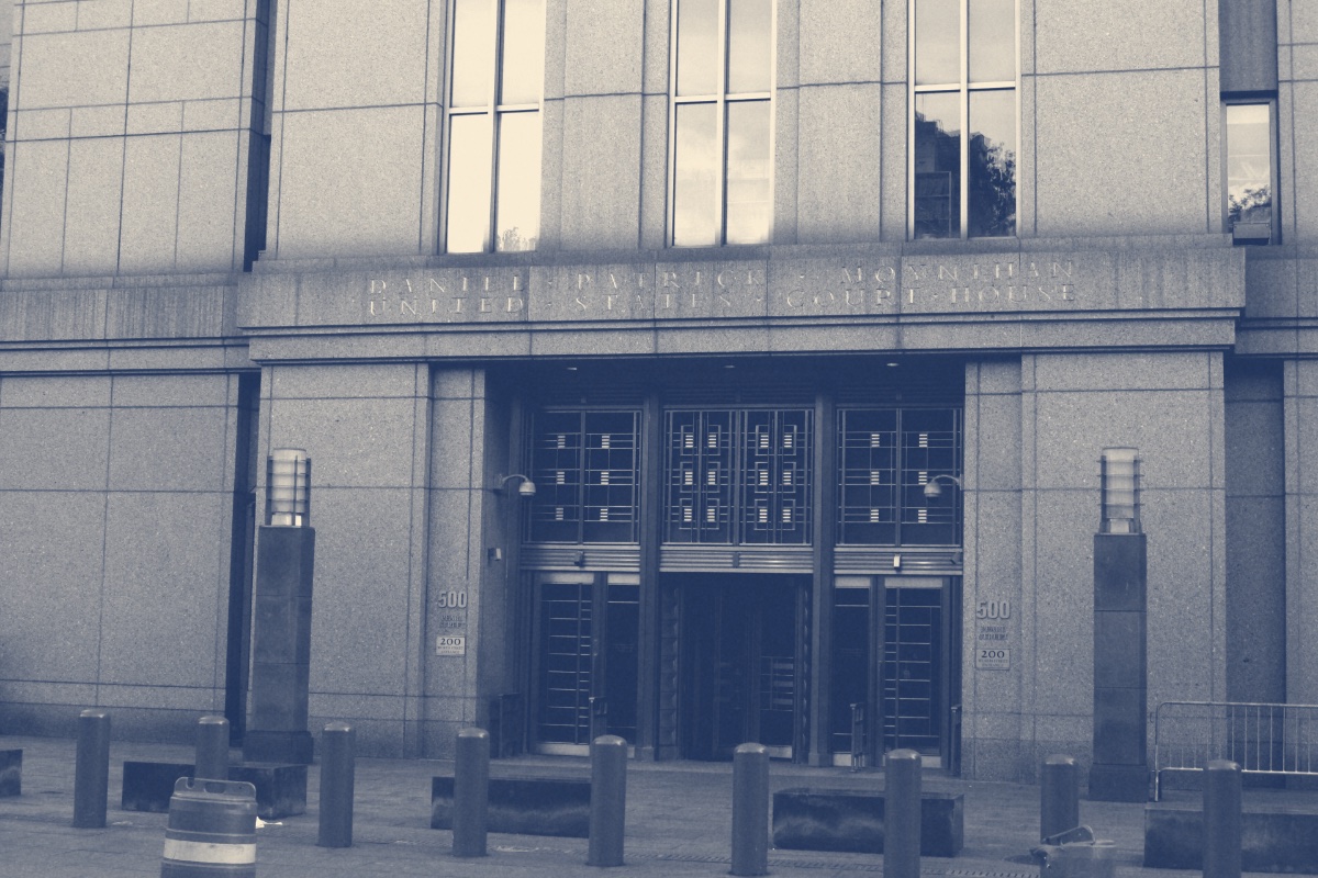 The entrance of the Daniel Patrick Moynihan United States Courthouse in Manhattan, its name carved in stone above tall bronze doors, bollards and two traffic barrels on the pavement in front, in a slate and bone duotone