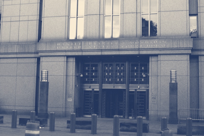 The entrance of the Daniel Patrick Moynihan United States Courthouse in Manhattan, its name carved in stone above tall bronze doors, bollards and two traffic barrels on the pavement in front, in a slate and bone duotone