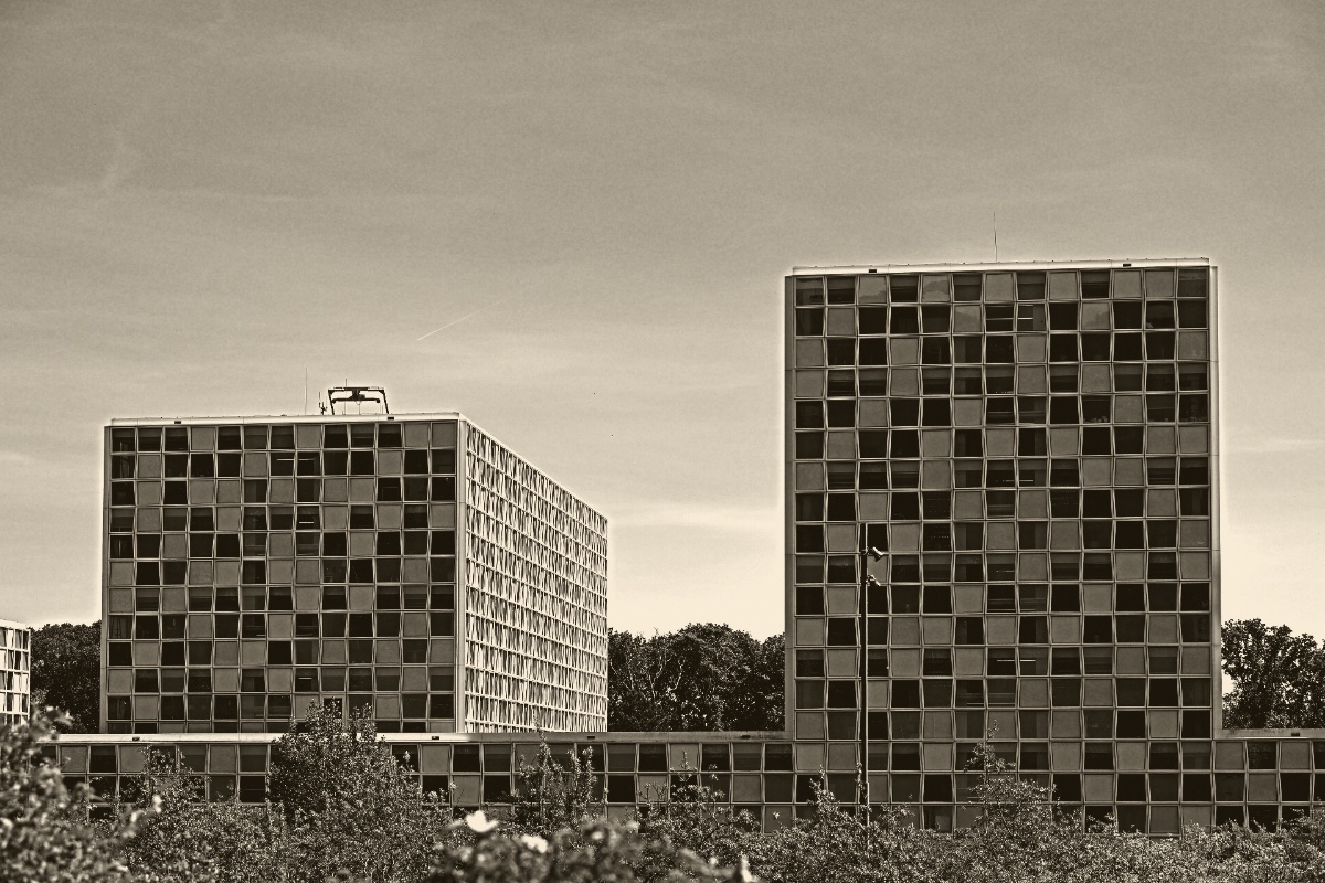 The International Criminal Court building in The Hague seen from across a green bank, two blocks of chequered glass panels rising above a low connecting wing under a clear blue sky, in an ink and bone duotone