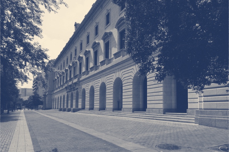 The John Minor Wisdom United States Court of Appeals Building in New Orleans, a long pale stone facade with tall arched ground-floor windows along a wide empty paved sidewalk, trees overhanging both sides, in a slate blue and bone duotone