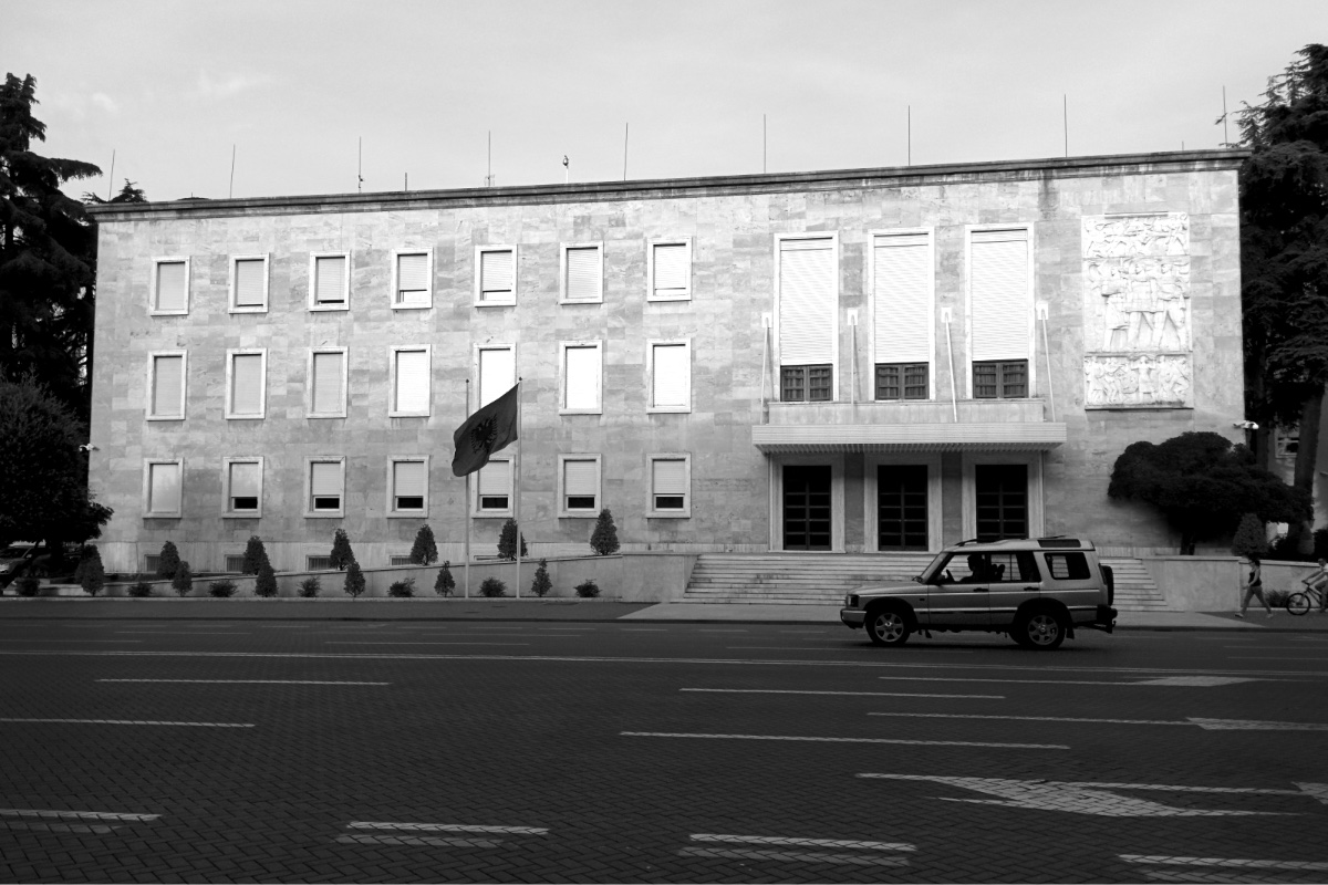 The office of the Prime Minister of Albania in Tirana, a long three-storey pale stone building with rows of shuttered windows, a carved relief panel at the right and a flag by the entrance, a parked four-wheel-drive car on the empty street in front, in black and white