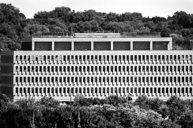 The Bishop Henry Whipple Federal Building near Fort Snelling, Minnesota, a long concrete block with rows of narrow windows above a band of trees, wooded hills behind, in grainy high-contrast black and white