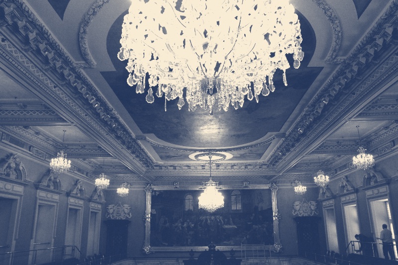 The Salon bleu of Quebec's National Assembly, a crystal chandelier hanging from a painted coffered ceiling above a large framed painting of a debate, in a slate and bone duotone