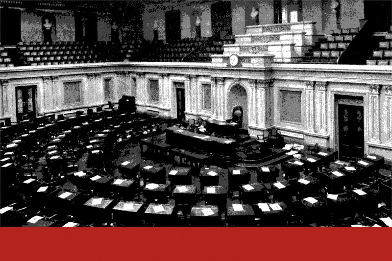 The Senate chamber photographed empty from the gallery, curved rows of wooden desks with papers laid on them facing the rostrum, with the public galleries above, in near black and white with a flat red band along the foot of the frame