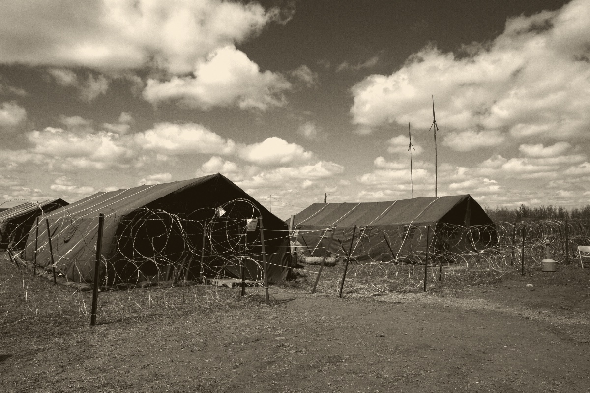 Two large olive military tents behind coils of razor wire at a field base camp on bare dusty ground, radio masts rising behind them under a sky full of heavy clouds, in a dark ink and bone duotone