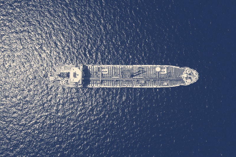 An oil tanker seen from directly above on dark open water, its long deck lined with pipework, in a slate and bone duotone