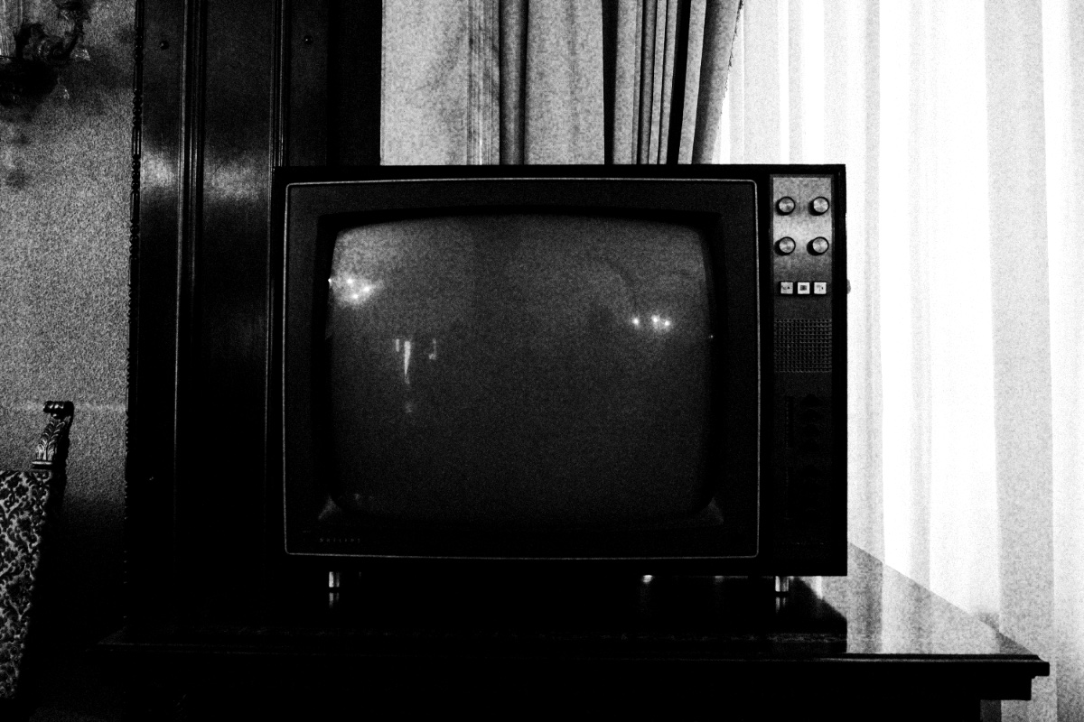 An old cathode-ray television set, switched off, on a dark wooden cabinet beside a curtained window, in high contrast black and white with coarse grain