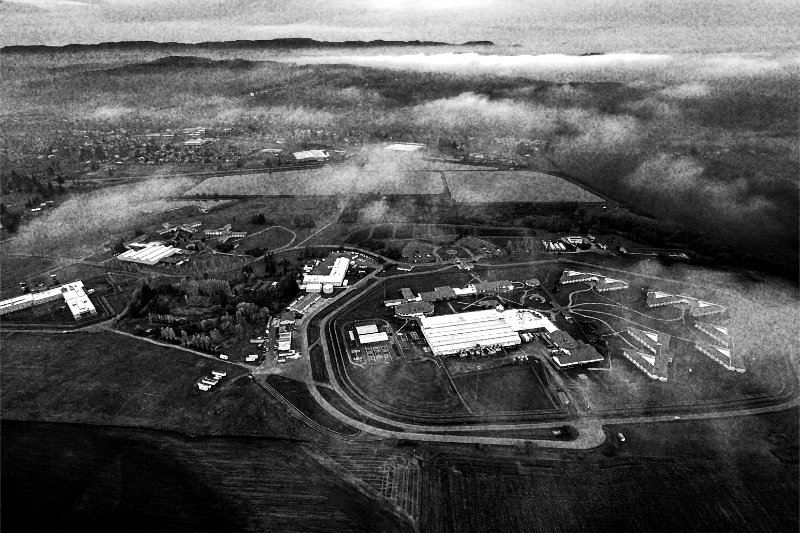 The Federal Correctional Institution at Sheridan, Oregon, seen from the air, low housing blocks and a large central building inside a looping perimeter road with fields and low cloud beyond, in high-contrast grainy black and white