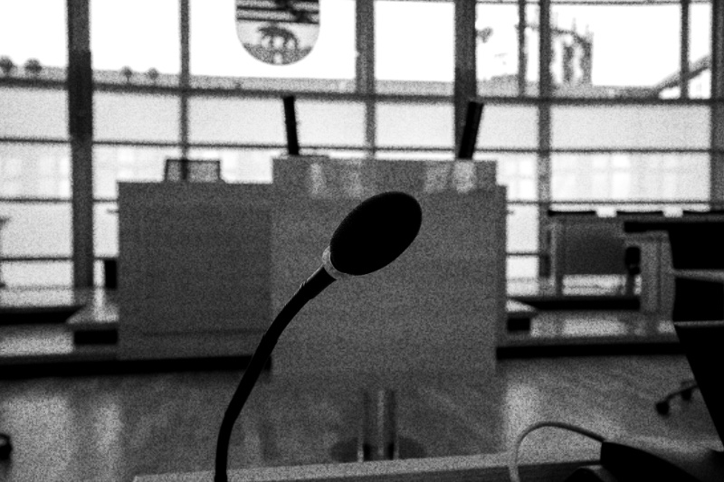 An empty speaker’s rostrum seen past a desk microphone in the plenary chamber of the Saxony-Anhalt state parliament, tall windows and the state coat of arms behind, in high contrast black and white with coarse grain