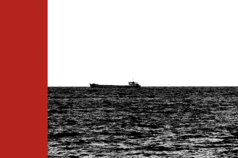 A single cargo ship alone on the horizon of an open sea, in near black and white with a flat red band down the left edge of the frame