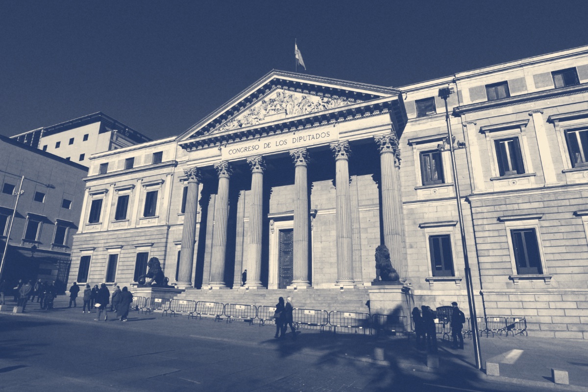 The columned portico of the Congress of Deputies in Madrid, its name carved above the columns and bronze lions at either side of the steps, pedestrians and crowd barriers in front, in a slate and bone duotone