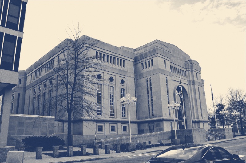 The Warren B. Rudman United States Courthouse in Concord, New Hampshire, a pale stone building with a tall arched entrance and rows of narrow windows behind a bare tree, a car passing in the street, in a slate blue and bone duotone