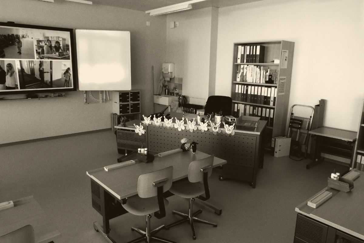 An empty primary school classroom with desks pushed together and children's work on the walls, in an ink and bone duotone