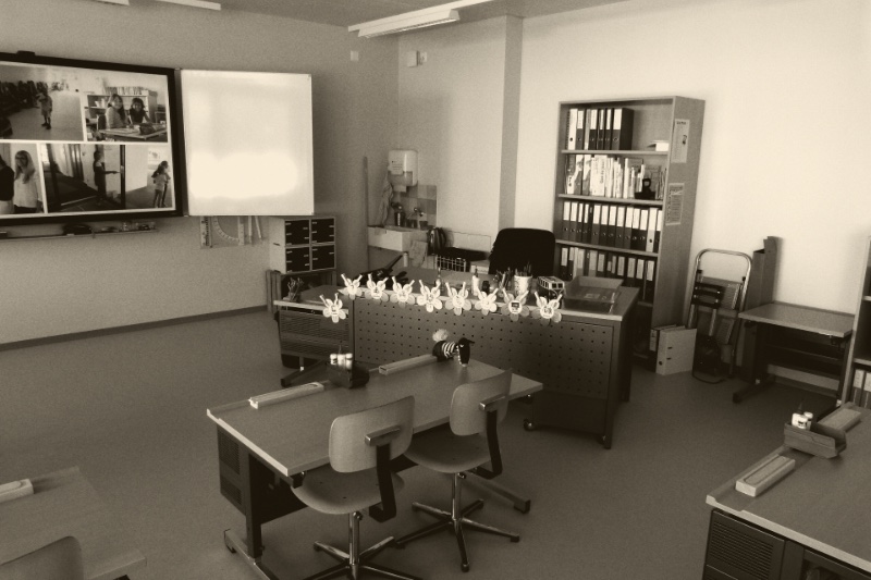 An empty primary school classroom with desks pushed together and children's work on the walls, in an ink and bone duotone