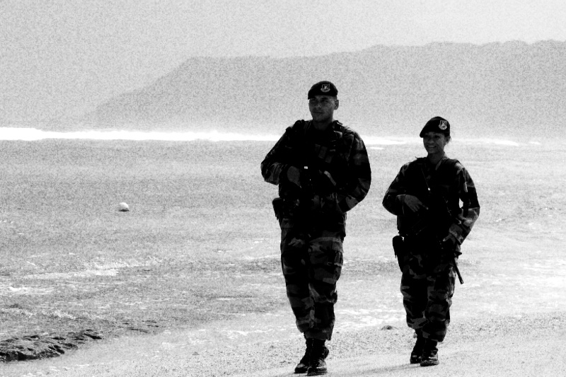 Two armed airmen walking along the sand at Tarague Beach in Guam, surf and a headland behind them, in high contrast black and white with coarse grain