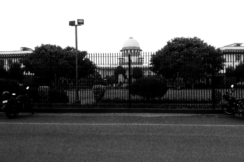 The Supreme Court of India in New Delhi, its dome visible above trees behind a tall iron fence, motorcycles parked on the road in front, in high contrast black and white with coarse grain