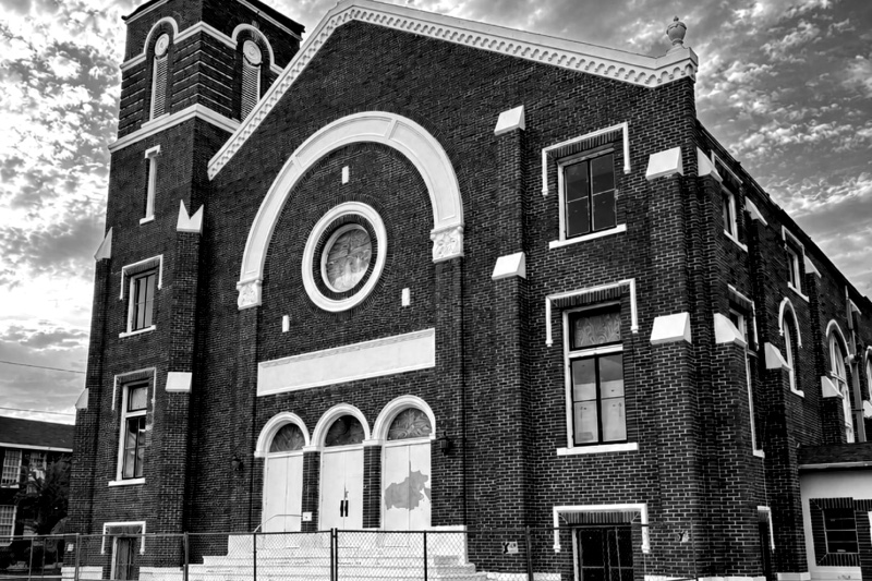 The red-brick facade of the former Euclid Methodist Church in St. Petersburg, Florida, with a white arched entrance, a round window and a square bell tower, a temporary chain-link fence across the front steps, in black and white
