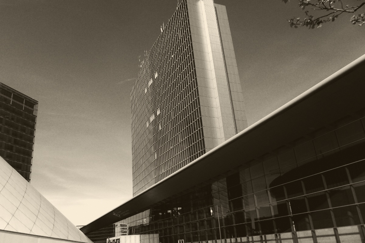 The European Convention Center in the Kirchberg district of Luxembourg, a long glass hall under a cantilevered roof with a tall office tower rising behind it, in a dark ink and bone duotone