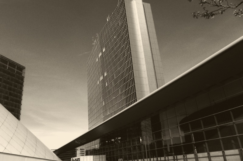 The European Convention Center in the Kirchberg district of Luxembourg, a long glass hall under a cantilevered roof with a tall office tower rising behind it, in a dark ink and bone duotone