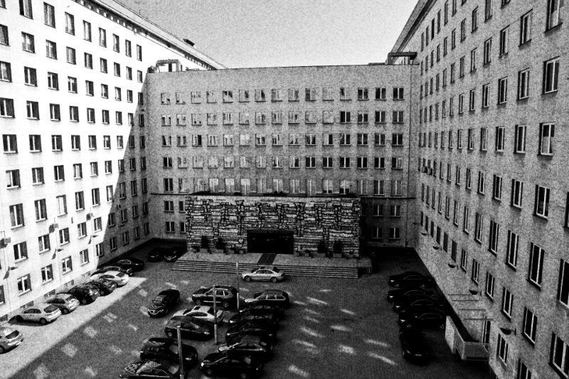 The courtyard of the National Bank of Poland headquarters in Warsaw seen from above, cars parked in rows between tall pale office wings, in high contrast black and white with coarse grain