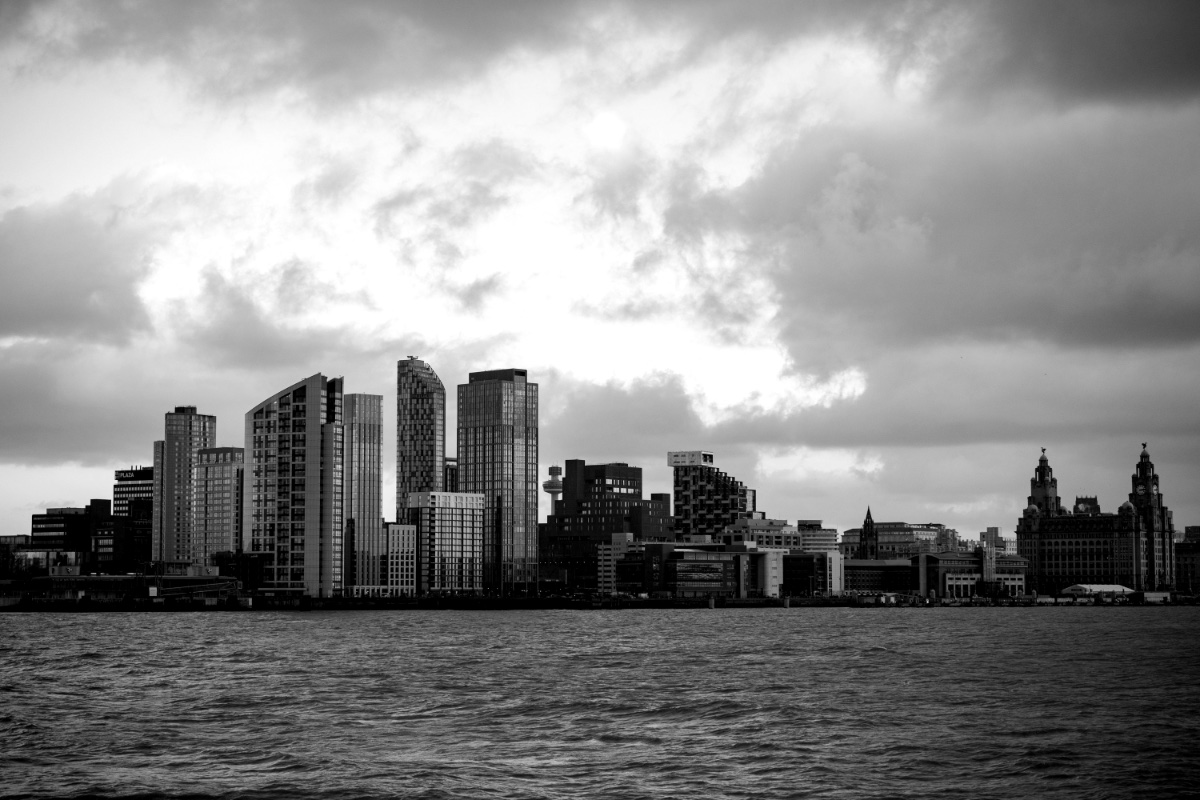 The Liverpool waterfront seen across the River Mersey under heavy cloud, towers and the Royal Liver Building along the far bank, in plain black and white