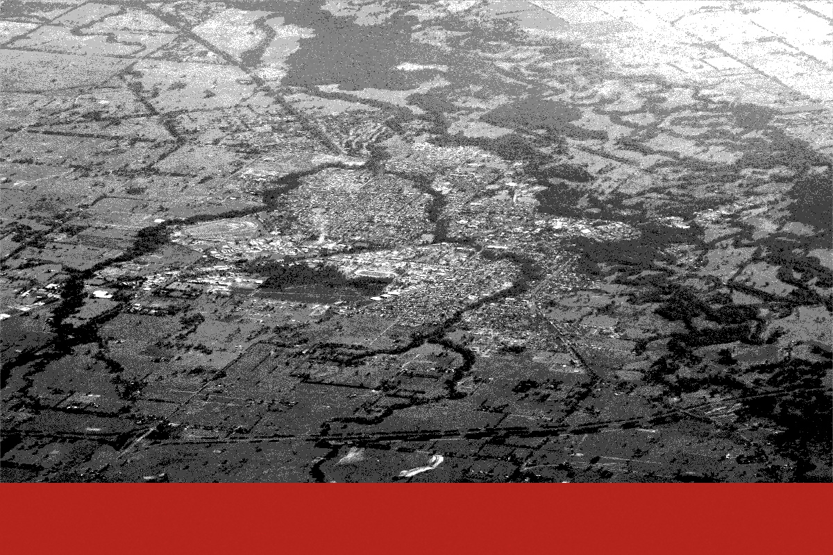 An aerial view of Wangaratta in north-east Victoria, the town spread across flat farmland with the river and its wooded flood channels winding through the paddocks, a broad flat red bar along the bottom edge, in near black and white