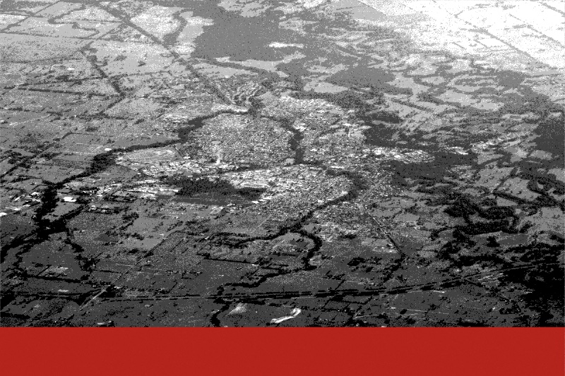 An aerial view of Wangaratta in north-east Victoria, the town spread across flat farmland with the river and its wooded flood channels winding through the paddocks, a broad flat red bar along the bottom edge, in near black and white