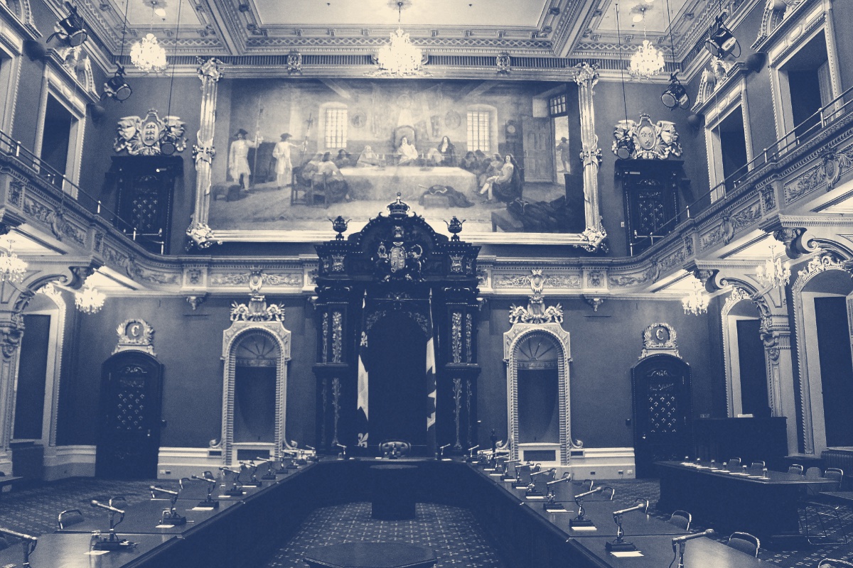 The empty Legislative Council chamber in the Quebec Parliament Building, ornate balconies around a carved wooden canopy topped by a crown, two flags hanging beside an empty chair, rows of desks with microphones in the foreground and a large historical painting above, in a slate blue and bone duotone