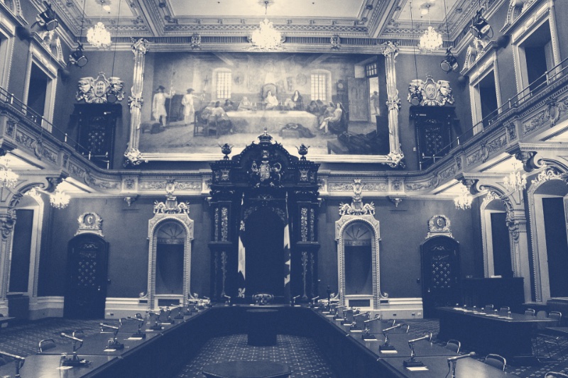 The empty Legislative Council chamber in the Quebec Parliament Building, ornate balconies around a carved wooden canopy topped by a crown, two flags hanging beside an empty chair, rows of desks with microphones in the foreground and a large historical painting above, in a slate blue and bone duotone
