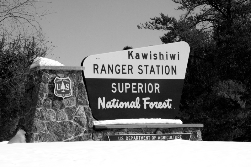 A brown and white sign reading Kawishiwi Ranger Station, Superior National Forest, with the Forest Service shield on its stone base, snow banked in front and pine trees behind, in black and white