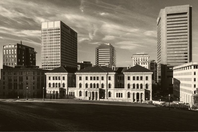 The Lewis F. Powell Jr. courthouse in Richmond, a long stone building with arched windows and red tile roofs across a sloping lawn, office towers rising behind it, in an ink and bone duotone