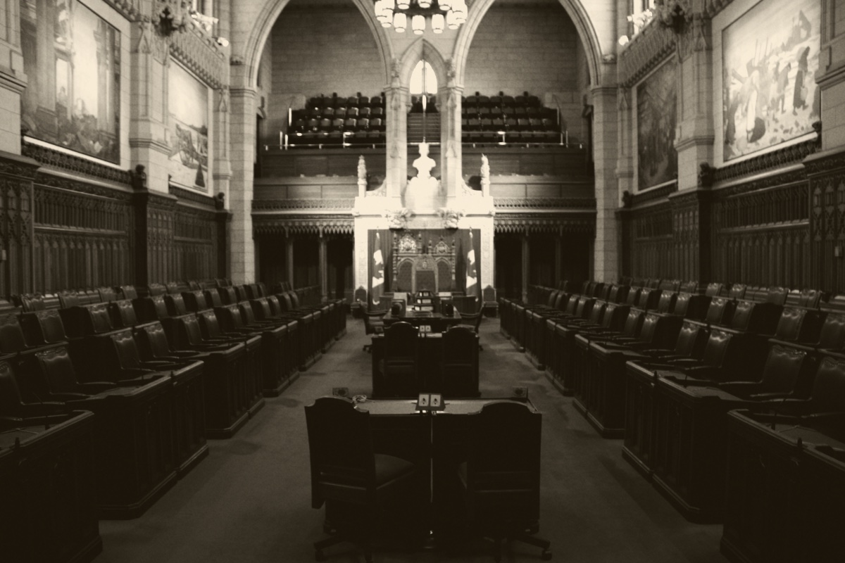The empty Senate chamber in the Centre Block of Parliament in Ottawa, rows of wooden desks and leather chairs on either side of a central aisle leading to the thrones under a stone arch, with large paintings on the walls, in a dark ink and bone duotone