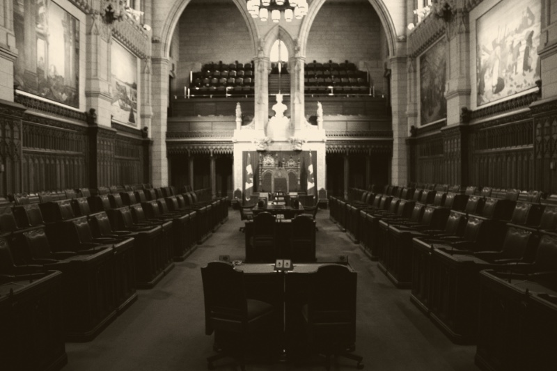 The empty Senate chamber in the Centre Block of Parliament in Ottawa, rows of wooden desks and leather chairs on either side of a central aisle leading to the thrones under a stone arch, with large paintings on the walls, in a dark ink and bone duotone