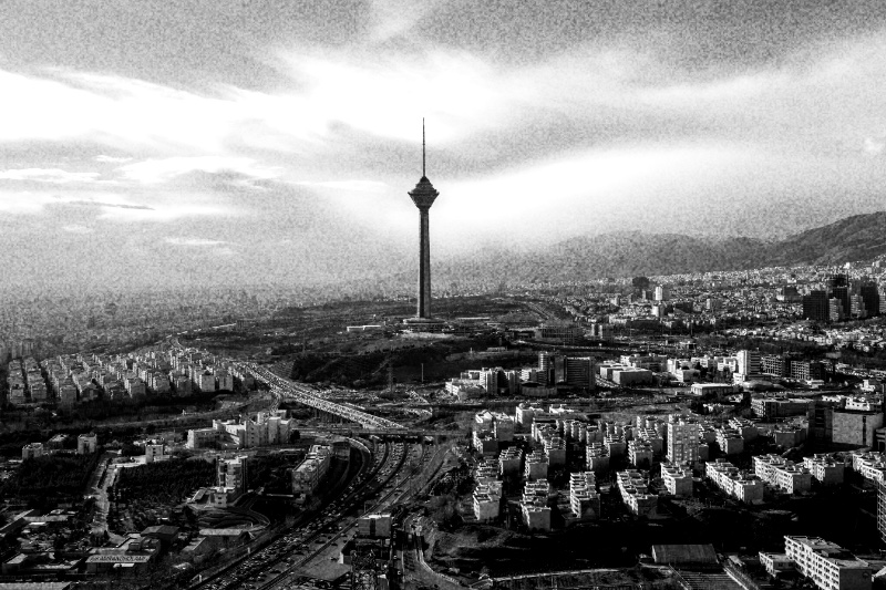 An aerial view of Tehran with the Milad Tower rising above the city and the Alborz mountains behind, in high contrast black and white with coarse grain