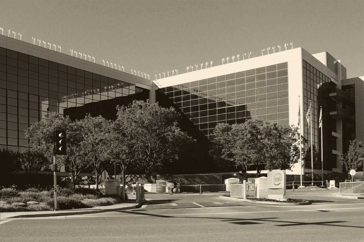 Intel's headquarters building in Santa Clara, a long glass curtain wall above a row of trees, with flagpoles, a gatehouse and a company sign at the entrance road, in a warm dark-brown and cream duotone