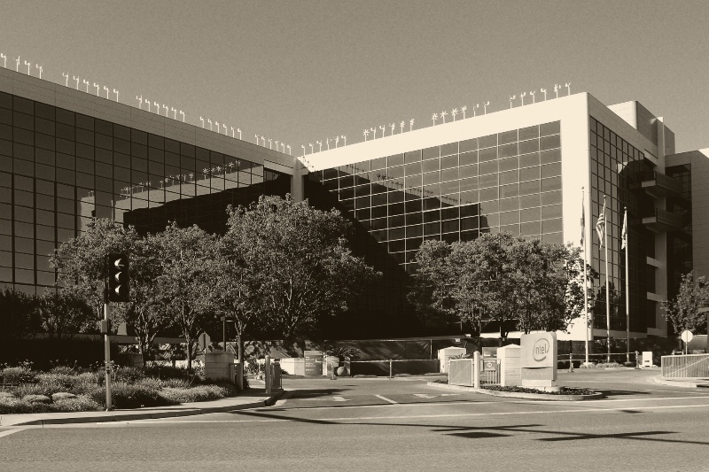 Intel's headquarters building in Santa Clara, a long glass curtain wall above a row of trees, with flagpoles, a gatehouse and a company sign at the entrance road, in a warm dark-brown and cream duotone
