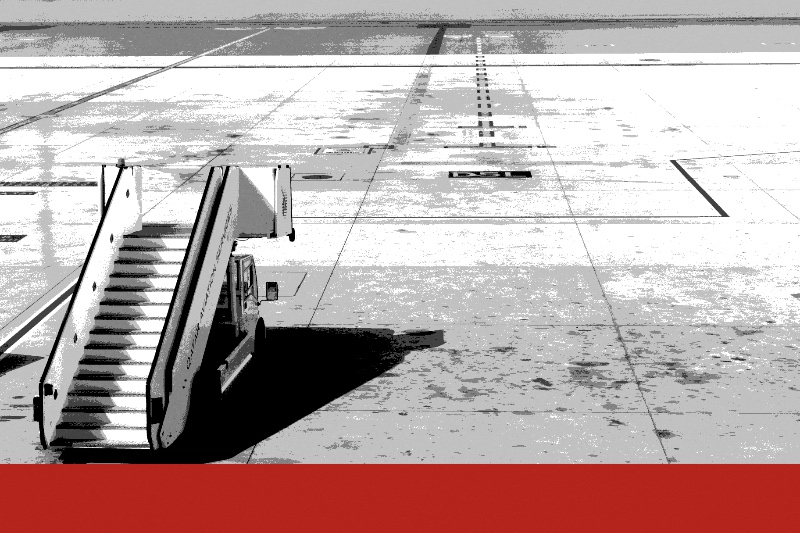 A set of mobile boarding stairs standing alone on an empty airport apron, in near black and white with a flat red band along the foot of the frame