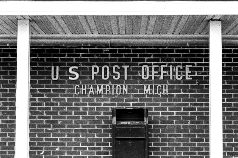 A blue United States Postal Service collection box beside a small rural post office, in high contrast black and white with coarse grain