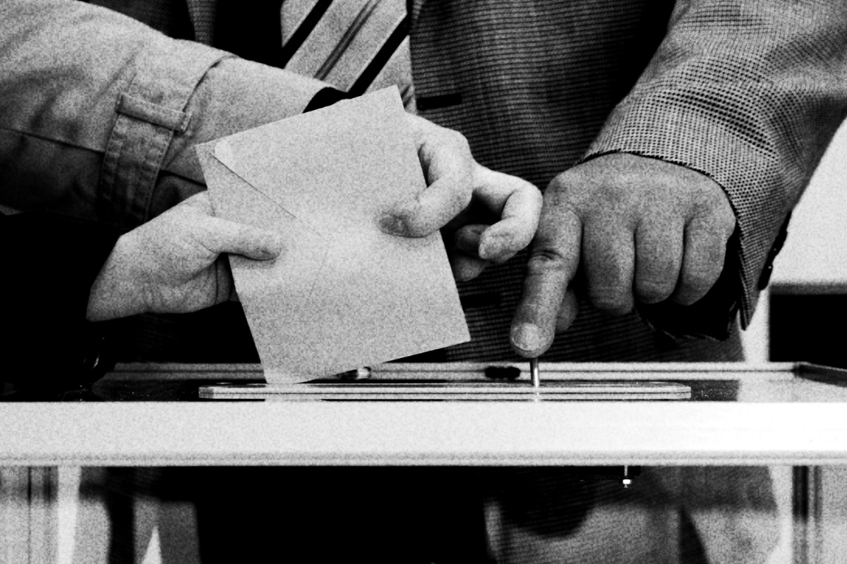 Two hands posting a brown envelope into the slot of a clear ballot box while a third hand points at the slot, in high contrast black and white with coarse grain