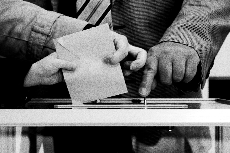 Two hands posting a brown envelope into the slot of a clear ballot box while a third hand points at the slot, in high contrast black and white with coarse grain