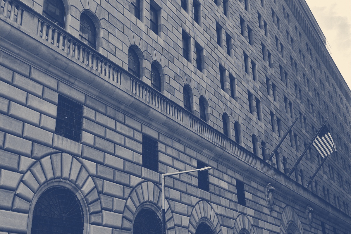 The stone facade of the Federal Reserve Bank of New York with arched windows and an American flag, in a slate and bone duotone