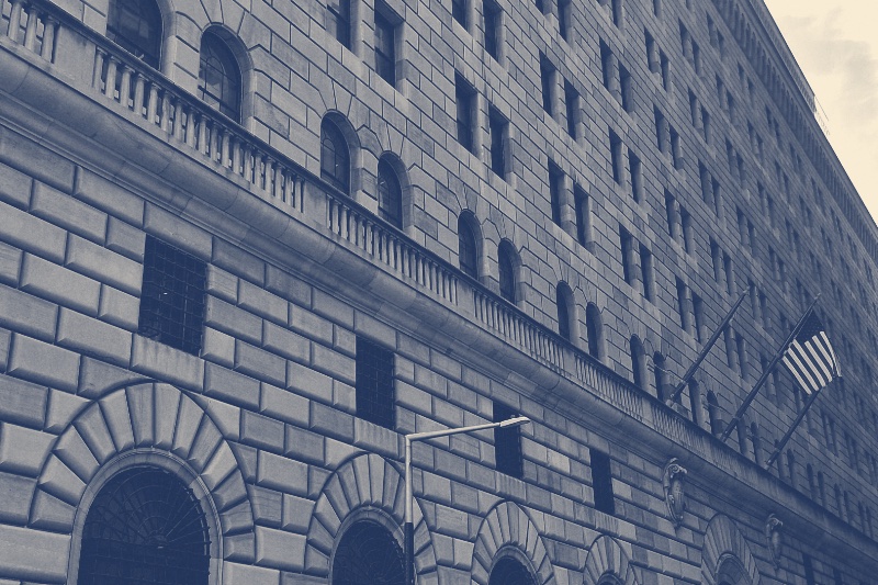 The stone facade of the Federal Reserve Bank of New York with arched windows and an American flag, in a slate and bone duotone