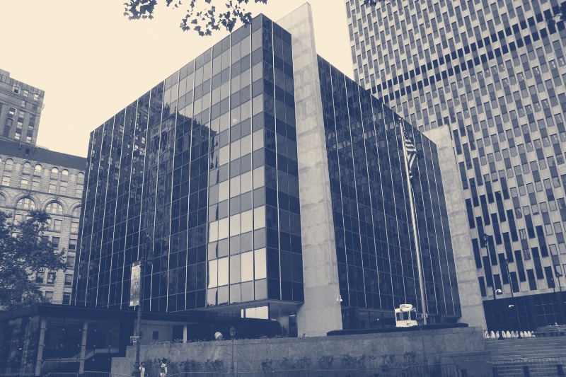 The glass-fronted building in New York that houses the Court of International Trade, a dark mirrored facade with a flag at its corner and a larger tower rising behind it, in a slate and bone duotone